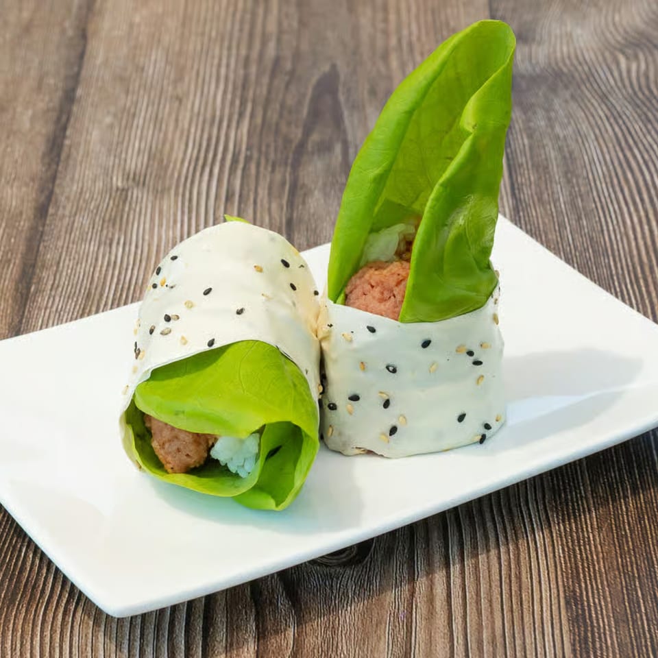 Two Higo Sushi rolls, wrapped in lettuce and rice paper with sesame seeds, on a white plate.